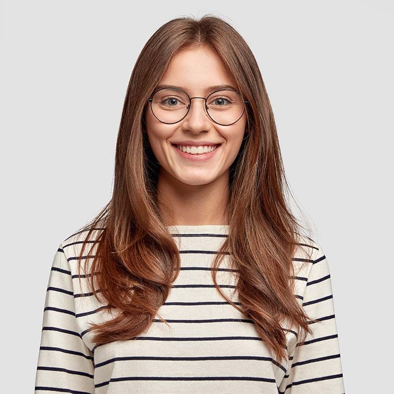 Indoor shot of attractive European schoolgirl has toothy smile, dressed in striped sweater, rejoices positive moment in life, wears round spectacles. People, lifestyle and positive emotions concept